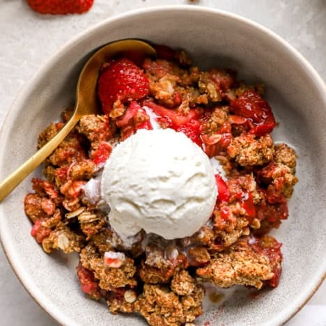 A bowl of strawberry crisp topped with vanilla ice cream with a spoon on the side.