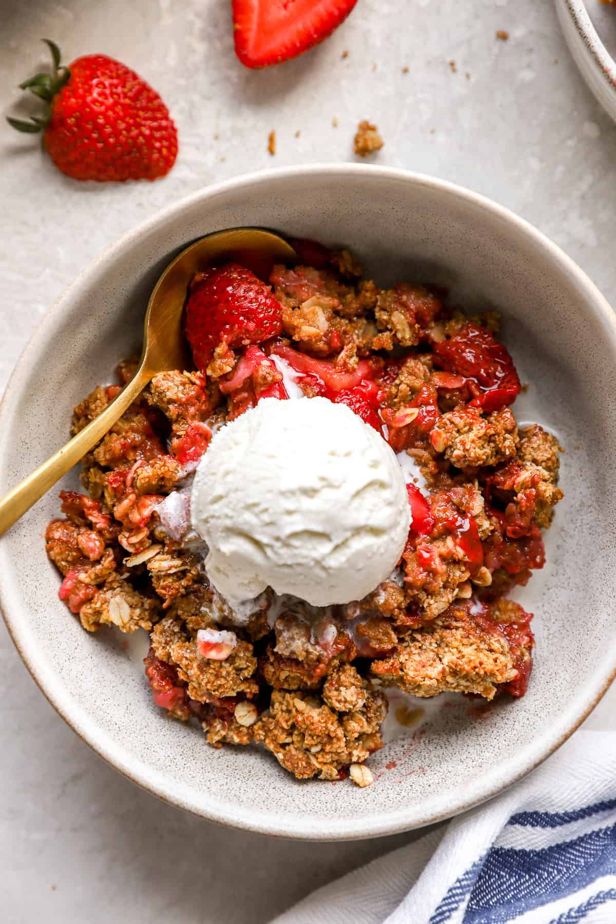 A bowl of strawberry crisp topped with vanilla ice cream with a spoon on the side.
