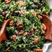 A fall kale salad with candied pecans, red onion, cheese, and shallot vinaigrette with two wooden serving spoons on top.