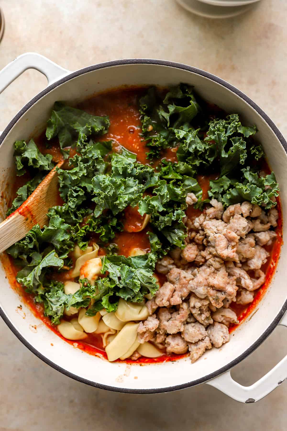 Kale, cooked sausage, and tortellini being added to a pot of soup. 
