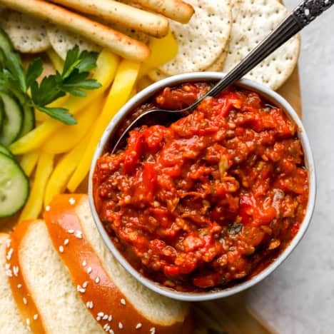 A bowl of matbucha with a spoon sticking out surrounded by veggies and crackers.