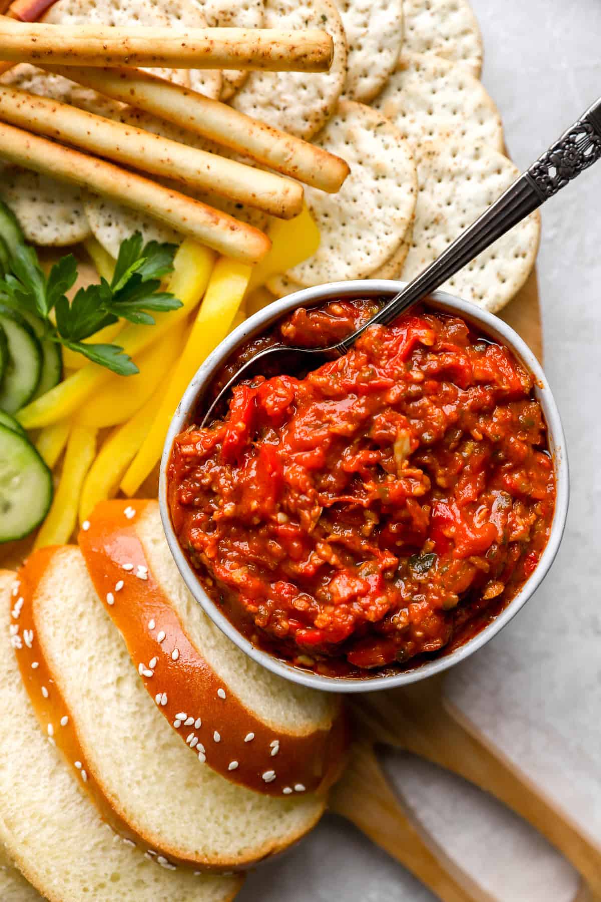 A bowl of matbucha with a spoon sticking out surrounded by veggies and crackers. 