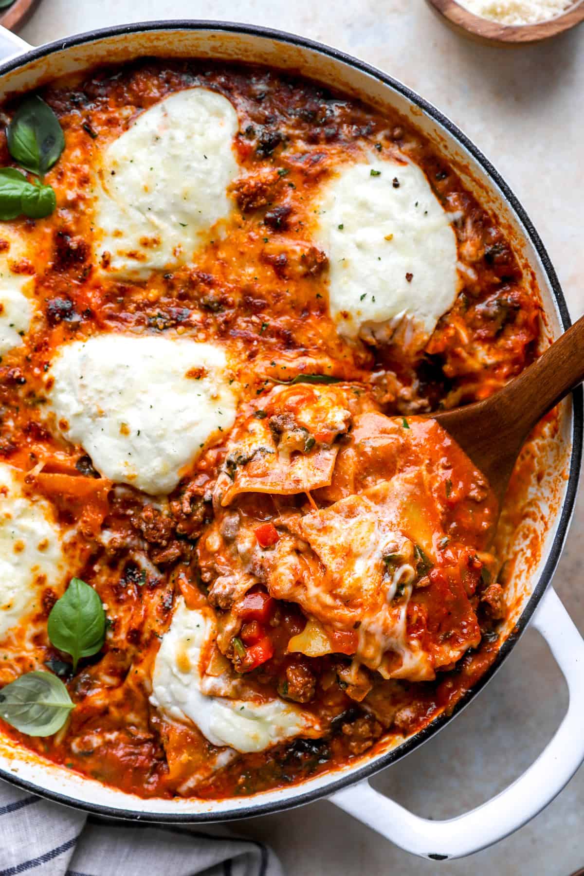 A wooden spoon lifting a scoop of lasagna from a skillet.