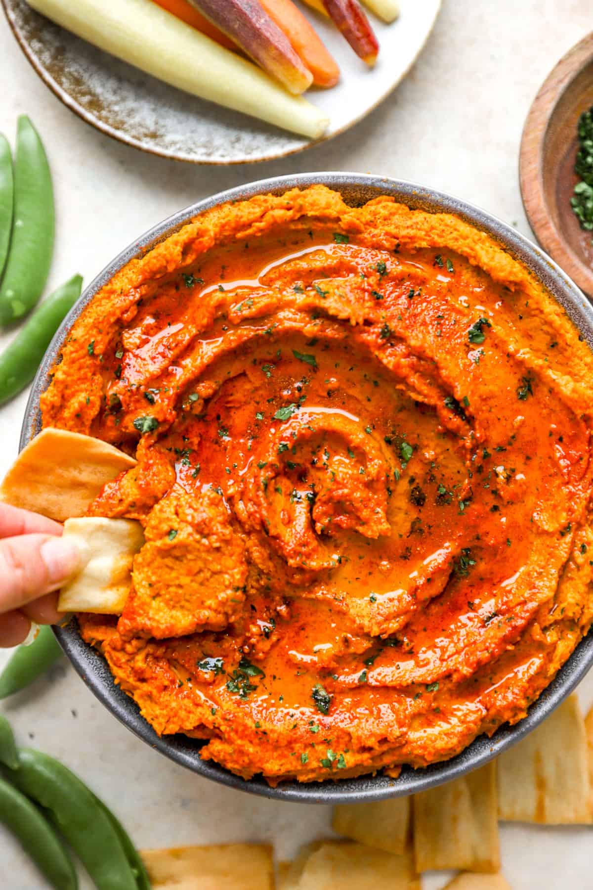 A hand dipping a cracker into a bowl of roasted carrot hummus.