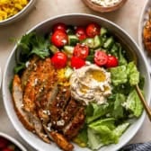 Sliced chicken cutlets on a salad with hummus, olive oil, and tomatoes.