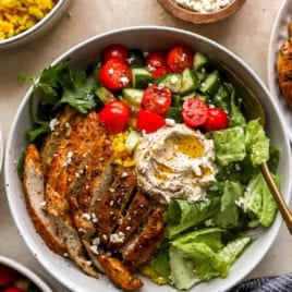 Sliced chicken cutlets on a salad with hummus, olive oil, and tomatoes.