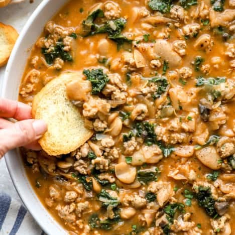 A hand dipping a piece of bread into a bowl of hot sausage Parmesan butter beans.