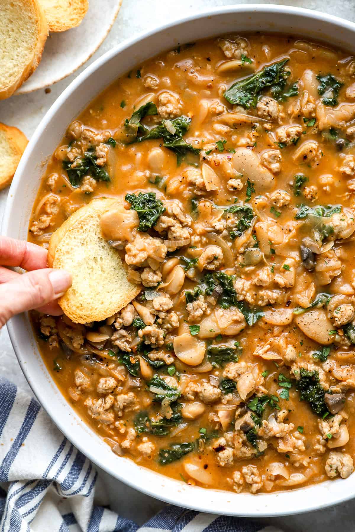 A hand dipping a piece of bread into a bowl of hot sausage Parmesan butter beans. 