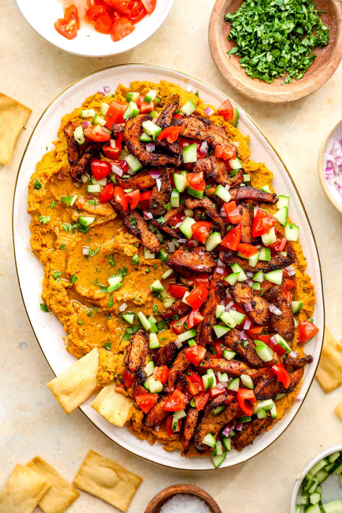 A loaded hummus platter topped with shawarma chicken, tomatoes, cucumbers, and feta.