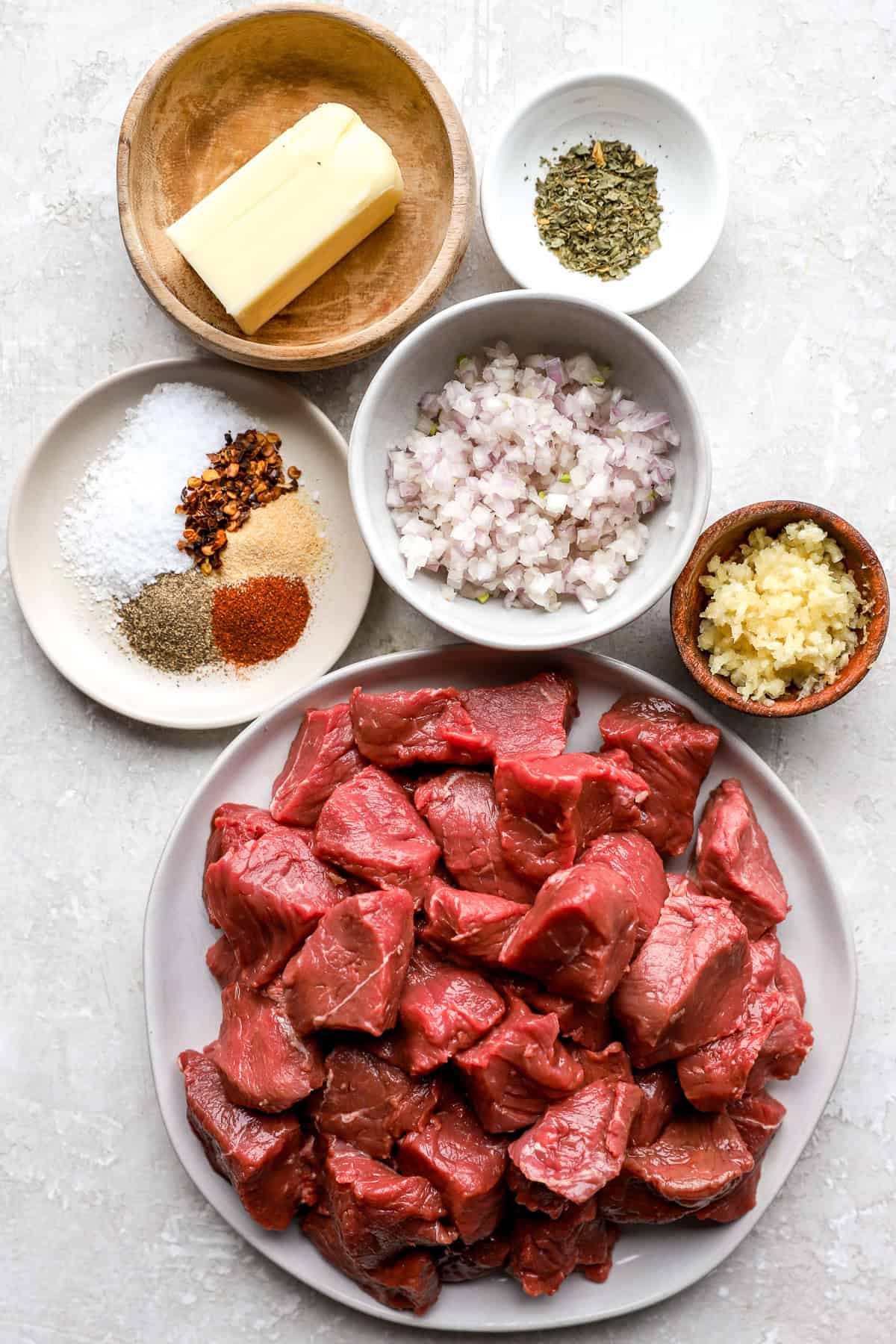 Butter, two bowls of spices, a bowl of garlic, a bowl of diced shallots, and a plate of raw steak bites.