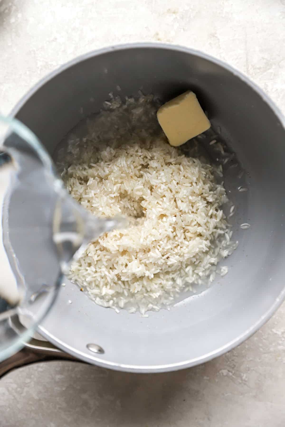 Dry white rice and butter in a pot, water being poured on top. 