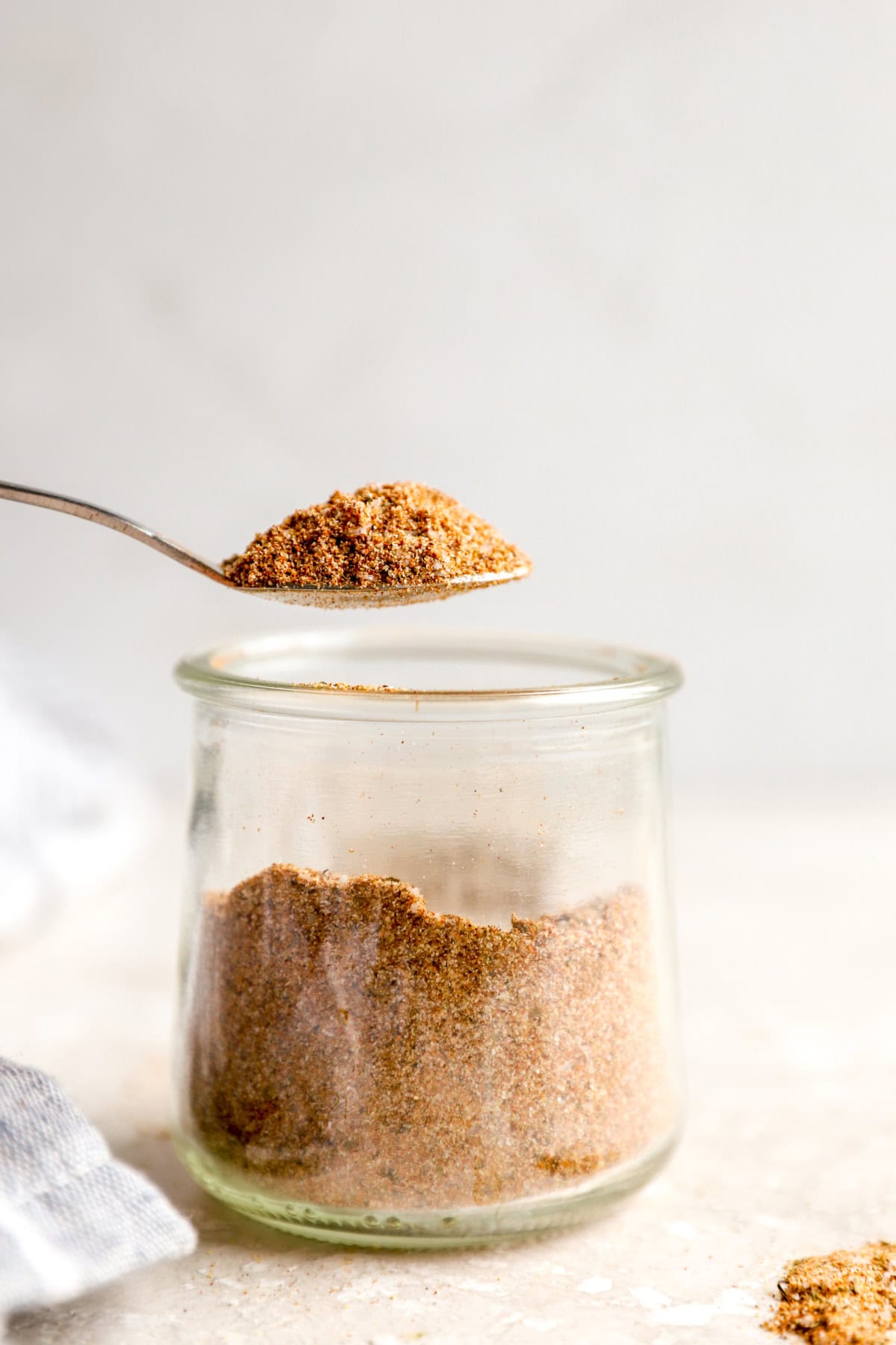 A spoon lifting a scoop of Sazón seasoning from a glass jar.
