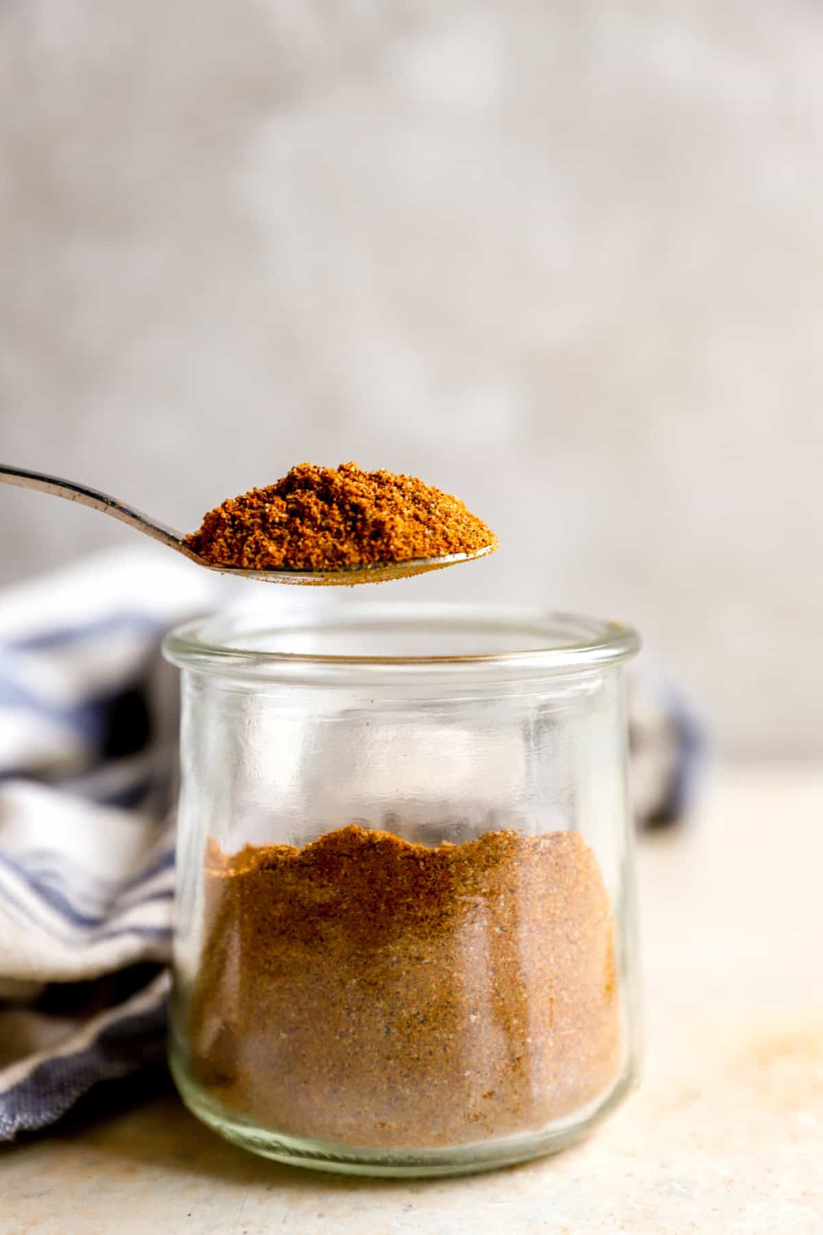 A spoon lifting a scoop of shawarma spice blend from a glass jar.