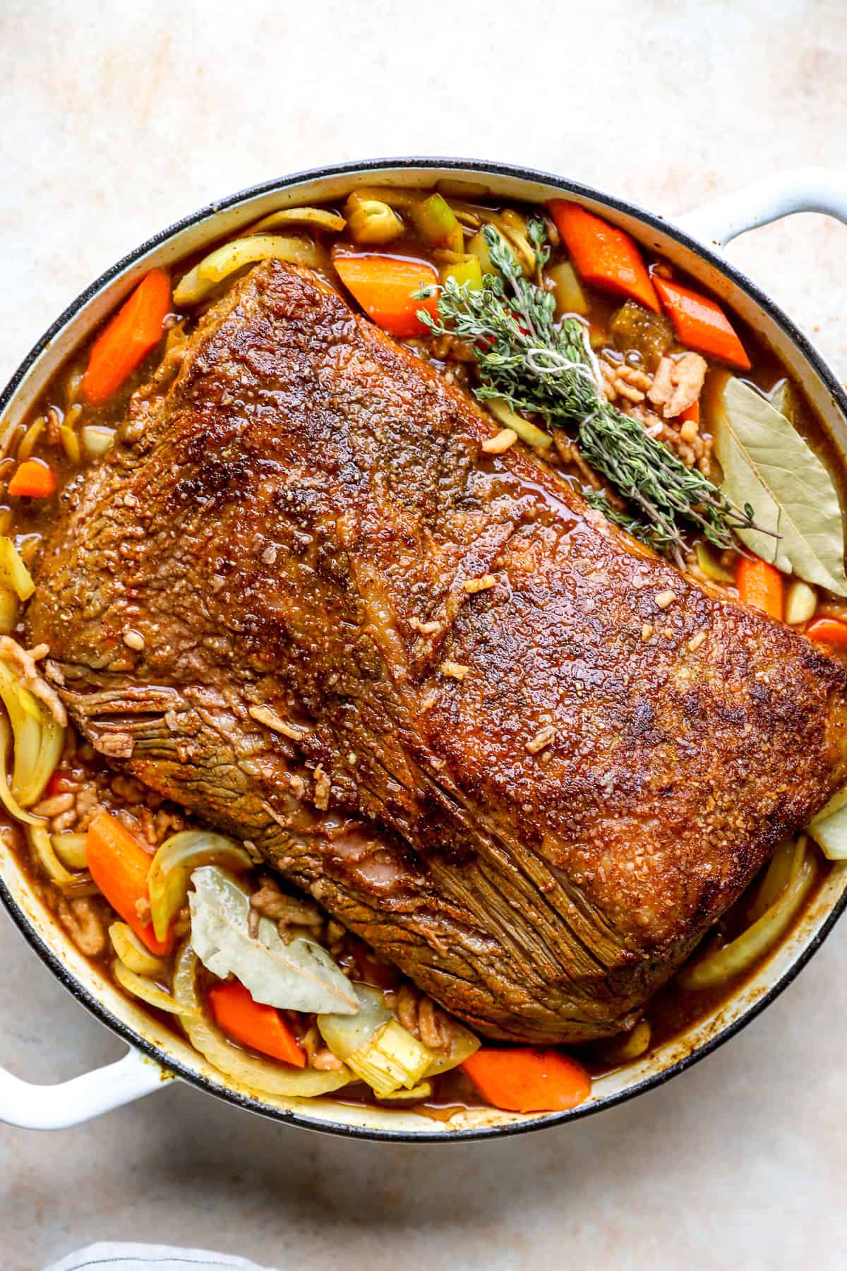 A brisket in a pot with carrots, onions, leeks, and herbs.