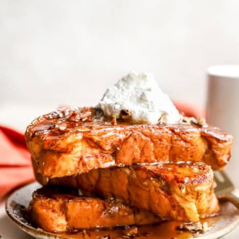 A stack of pumpkin French toast on a plate topped with whipped cream, powdered sugar, and maple syrup.